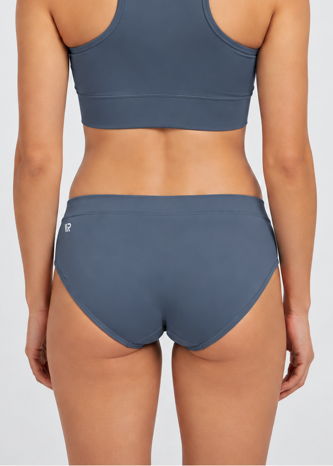 A woman viewed from the back wearing a matching slate blue-gray athletic set consisting of a sports bra and high-waisted briefs, with a small white logo on the back of the sports bra.