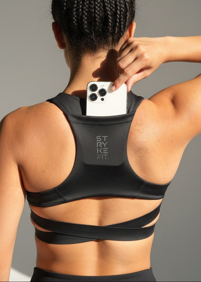 A woman modeling a black athletic sports bra and matching high-waisted shorts against a solid gray studio background. The brand name 'STRYKE FIT' is printed in reflective on the back of the sports bra. The ATHLETE CROP TOP features a discrete back pocket, keeping your essentials close, whether it's your phone, gels, keys, or anything you need to go the distance without distractions.

The performance racerback design follows the natural line of your shoulders, allowing unrestricted movement