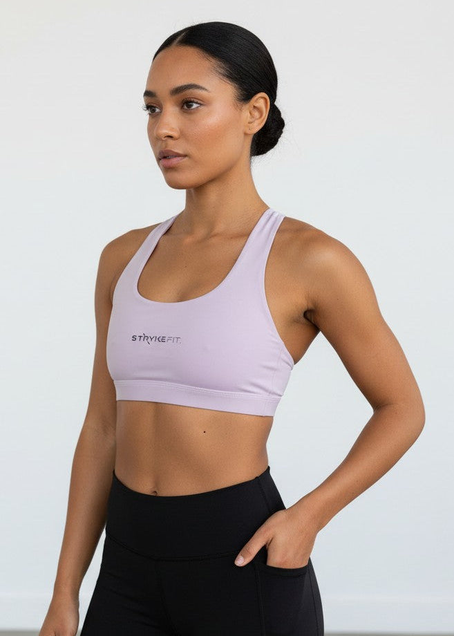 A woman modeling a light lavender sports bra and black high-waisted biker shorts against a plain white studio background. The ATHLETE CROP TOP features a discrete back pocket, keeping your essentials close, whether it's your phone, gels, keys, or anything you need to go the distance without distractions.

The performance racerback design follows the natural line of your shoulders, allowing unrestricted movement and complete freedom through every stride. Engineered for support without compromising comfort