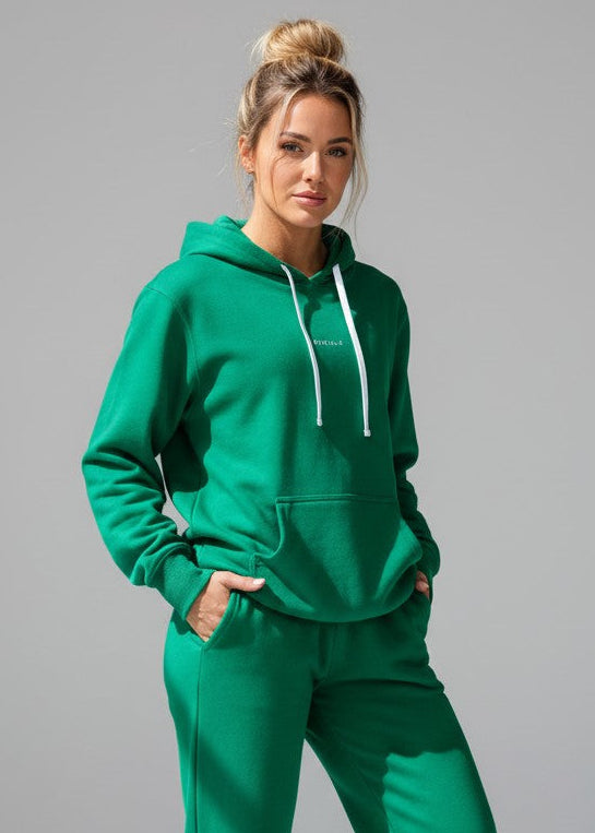 Woman wearing a green hoodie and shorts set on a white background. The Core Hooded Fleece is designed for everyday wear, combining comfort with effortless style. Made from premium brushed-back fleece, it delivers a soft, warm feel while remaining easy to wear and versatile. DETAILS, Premium brushed fleece, Self-lined hood, Kangaroo pocket, Soft ribbed cuffs, Printed logo